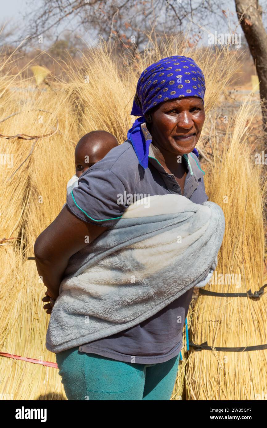 Femme africaine portant le bébé dans le dos enveloppé dans une serviette de la manière traditionnelle, chaume herbe en arrière-plan, la vie du village. Banque D'Images