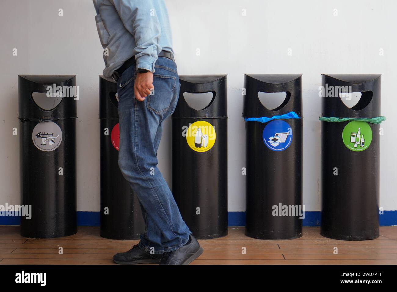 Angle frontal, capture une vue partielle d'un homme adulte debout à côté de quatre bacs de recyclage à code couleur dans un environnement intérieur. L'homme, habillé dans un l Banque D'Images