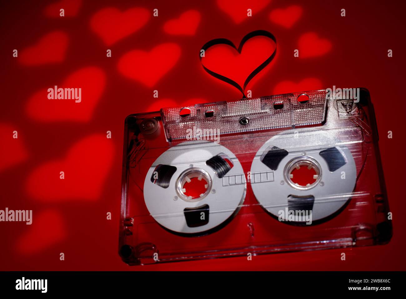 Cassette audio transparente avec ruban en forme de coeur à l'extérieur et avec beaucoup de projections de coeur de lumière rouge dans le fond rouge foncé. Banque D'Images