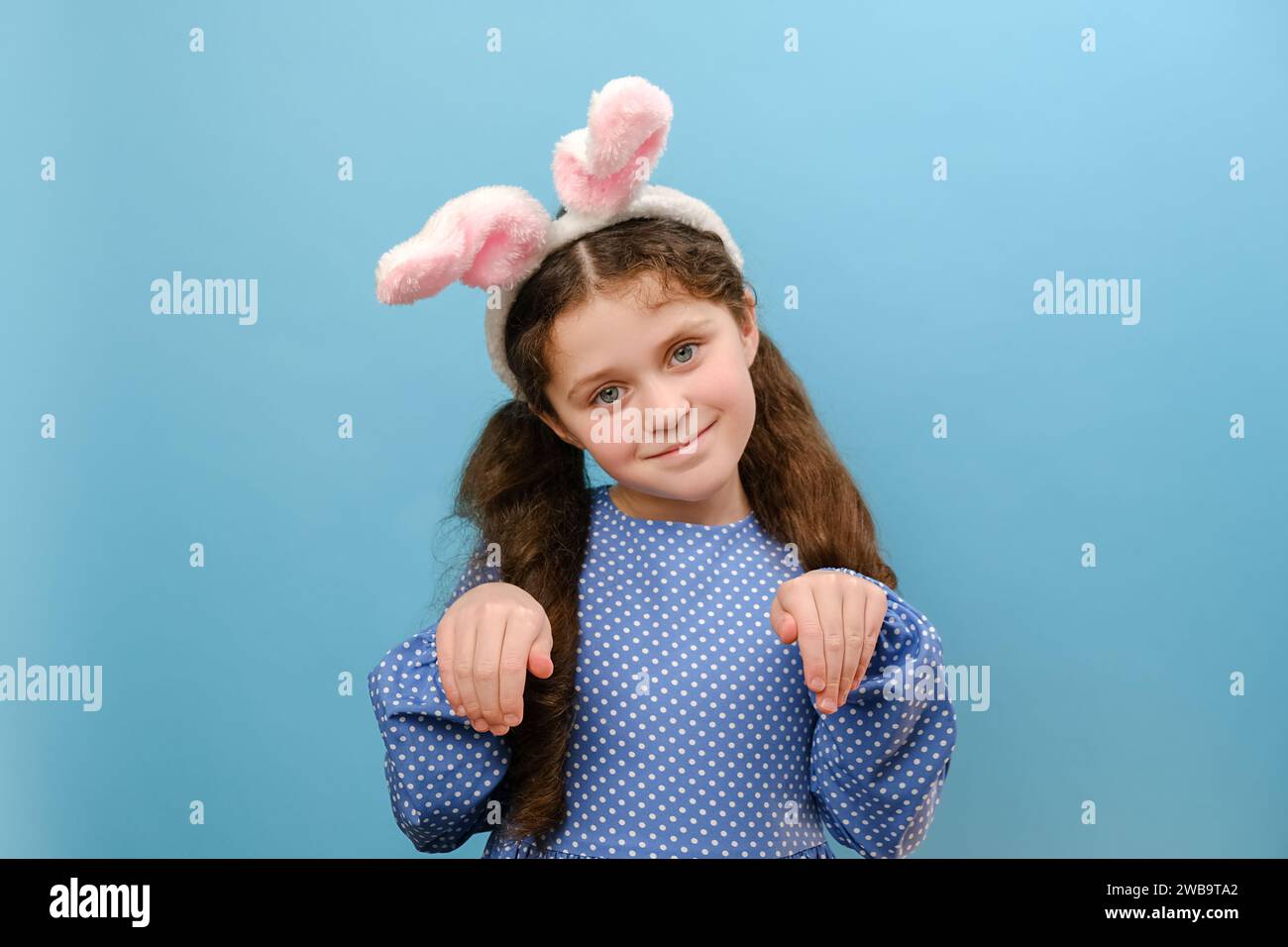 Portrait de drôle de petite fille ludique enfant portant rose pelucheux oreilles lapin les bras font des pattes, heureux regardant la caméra, debout isolé sur la couleur bleue Banque D'Images