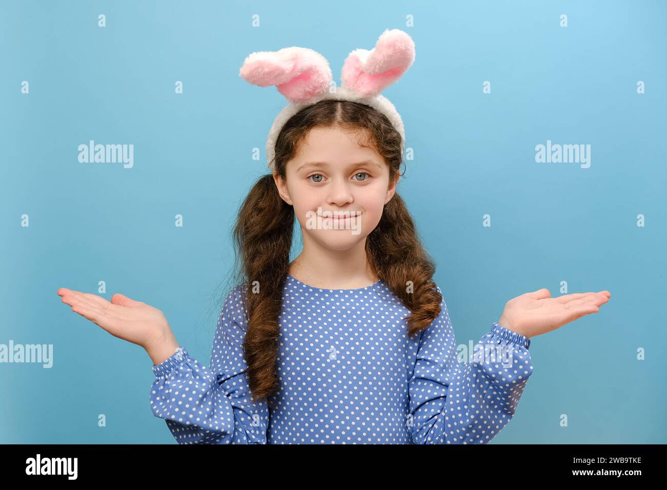 Portrait de jolie petite fille enfant dans les oreilles pelucheuses de lapin rose tient l'espace de copie avec des paumes, a la bonne humeur, posant sur le mur de fond de studio bleu avec em Banque D'Images