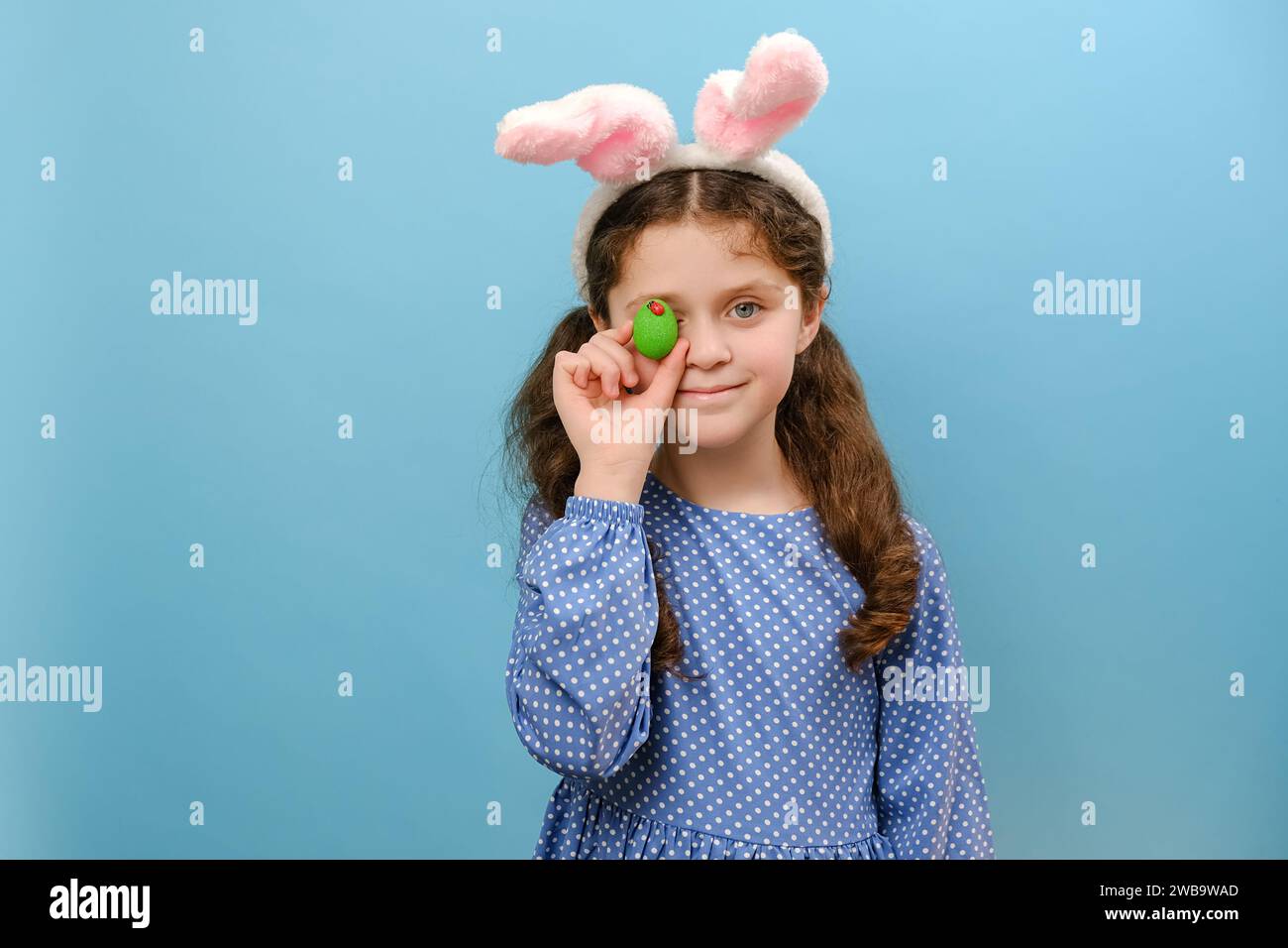 Portrait d'un gamin de préteen drôle et joyeux portant des oreilles de lapin rose de pâques moelleuses, tenant près du visage vert oeuf de Pâques, posant isolé sur le bleu Banque D'Images