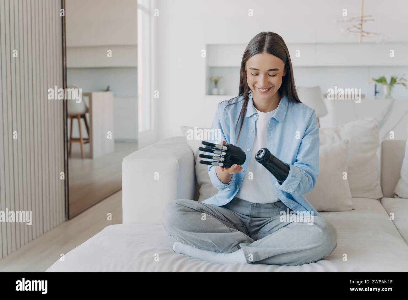 Joyeuse jeune femme avec un bras bionique assis sur un canapé, explorant la fonctionnalité de son membre prothétique dans une pièce lumineuse Banque D'Images