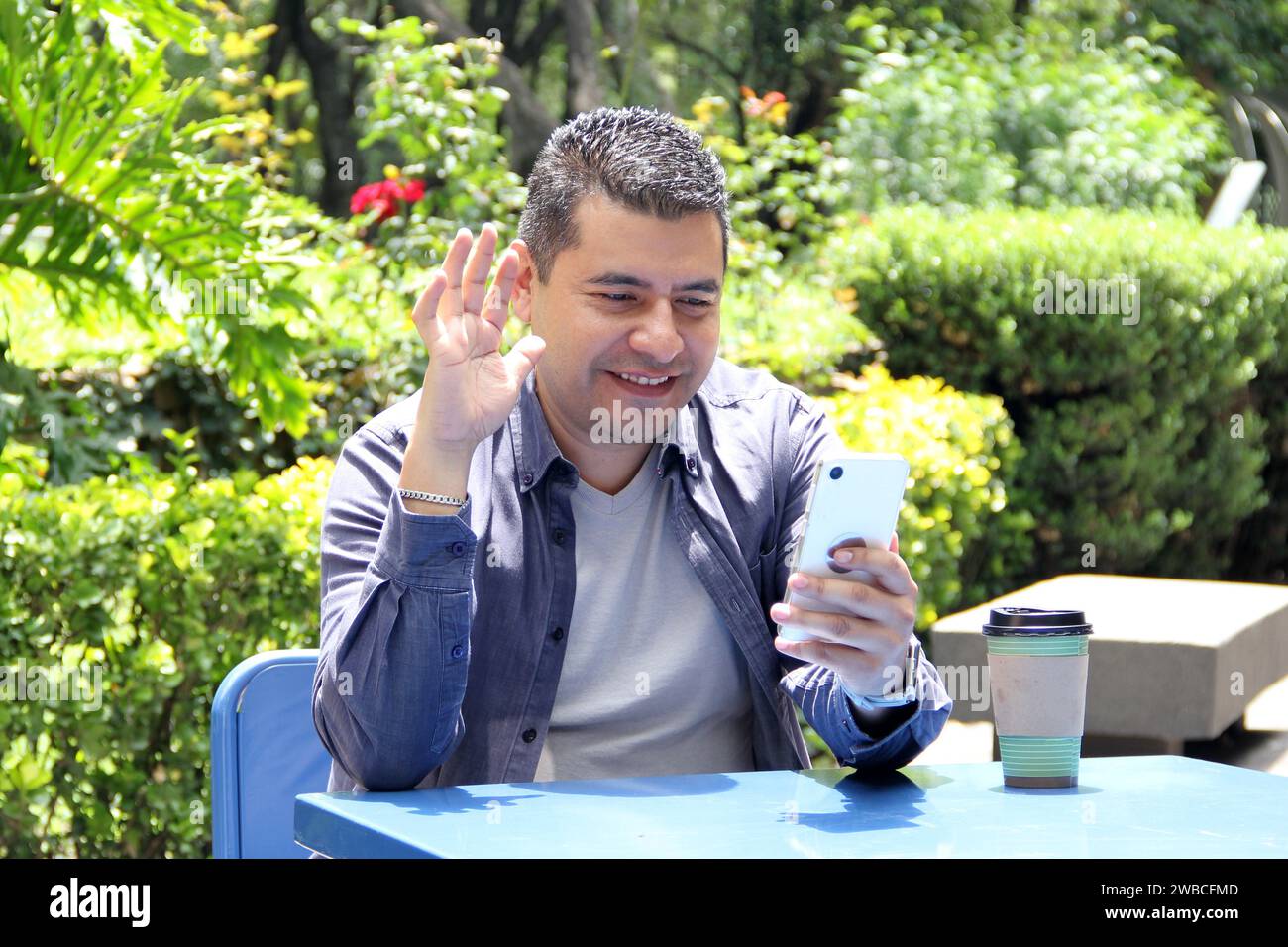 Un Latino de 40 ans à la peau foncée boit un café et utilise son téléphone portable sur un banc du parc pour travailler à distance, passer des appels, envoyer des messages, acheter et payer Banque D'Images