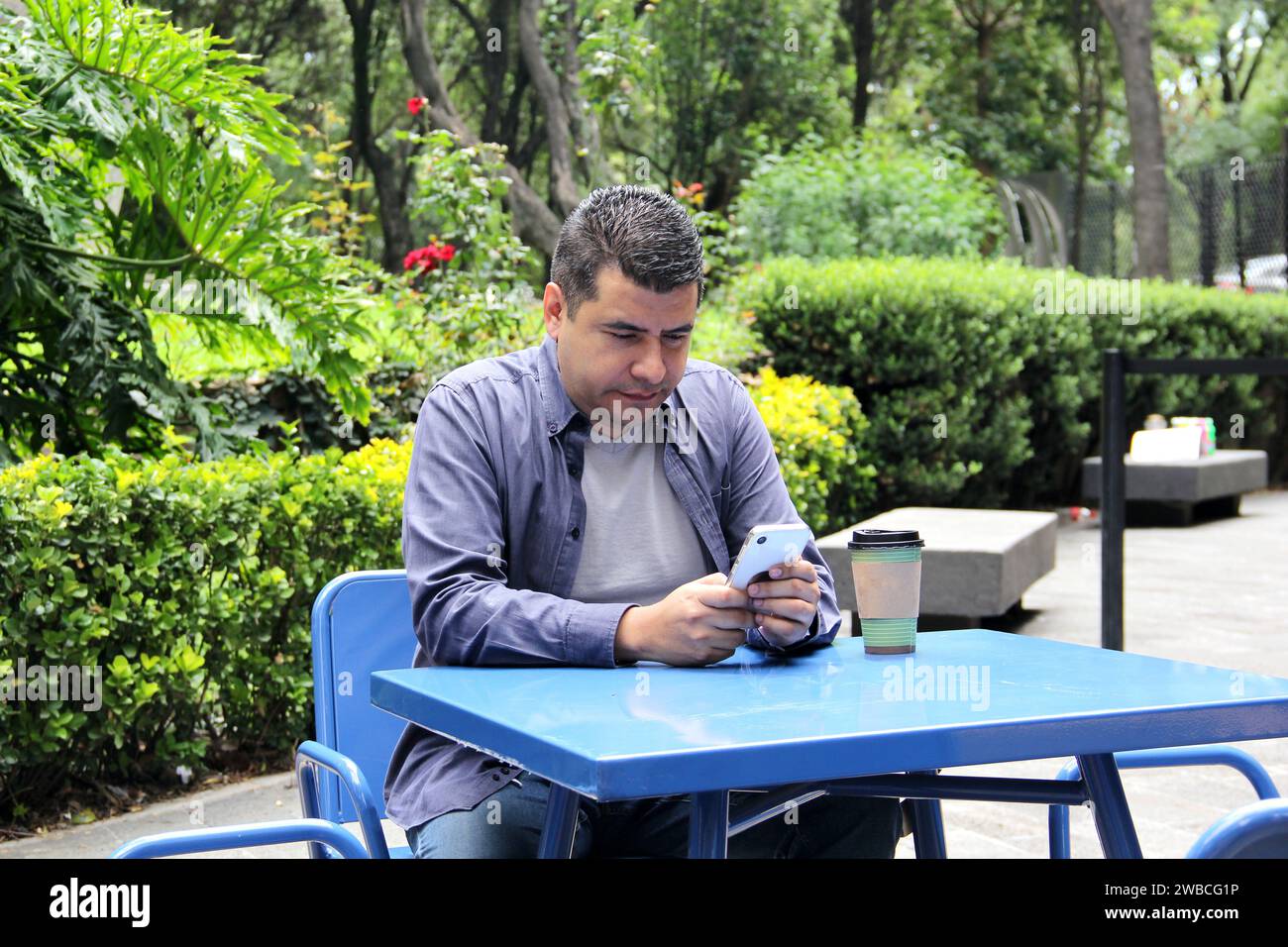 Un Latino de 40 ans à la peau foncée boit un café et utilise son téléphone portable sur un banc du parc pour travailler à distance, passer des appels, envoyer des messages, acheter et payer Banque D'Images