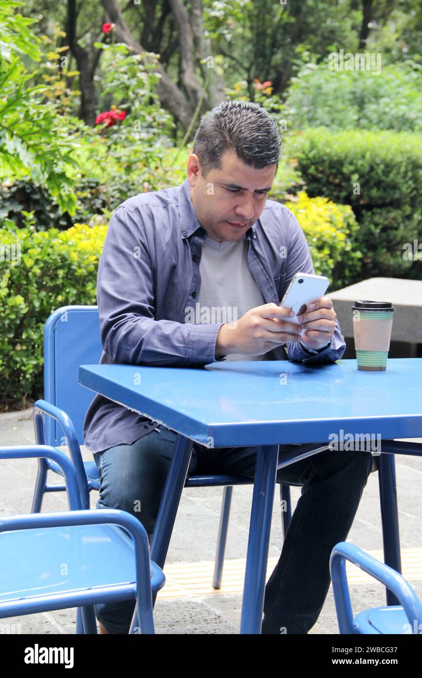Un Latino de 40 ans à la peau foncée boit un café et utilise son téléphone portable sur un banc du parc pour travailler à distance, passer des appels, envoyer des messages, acheter et payer Banque D'Images
