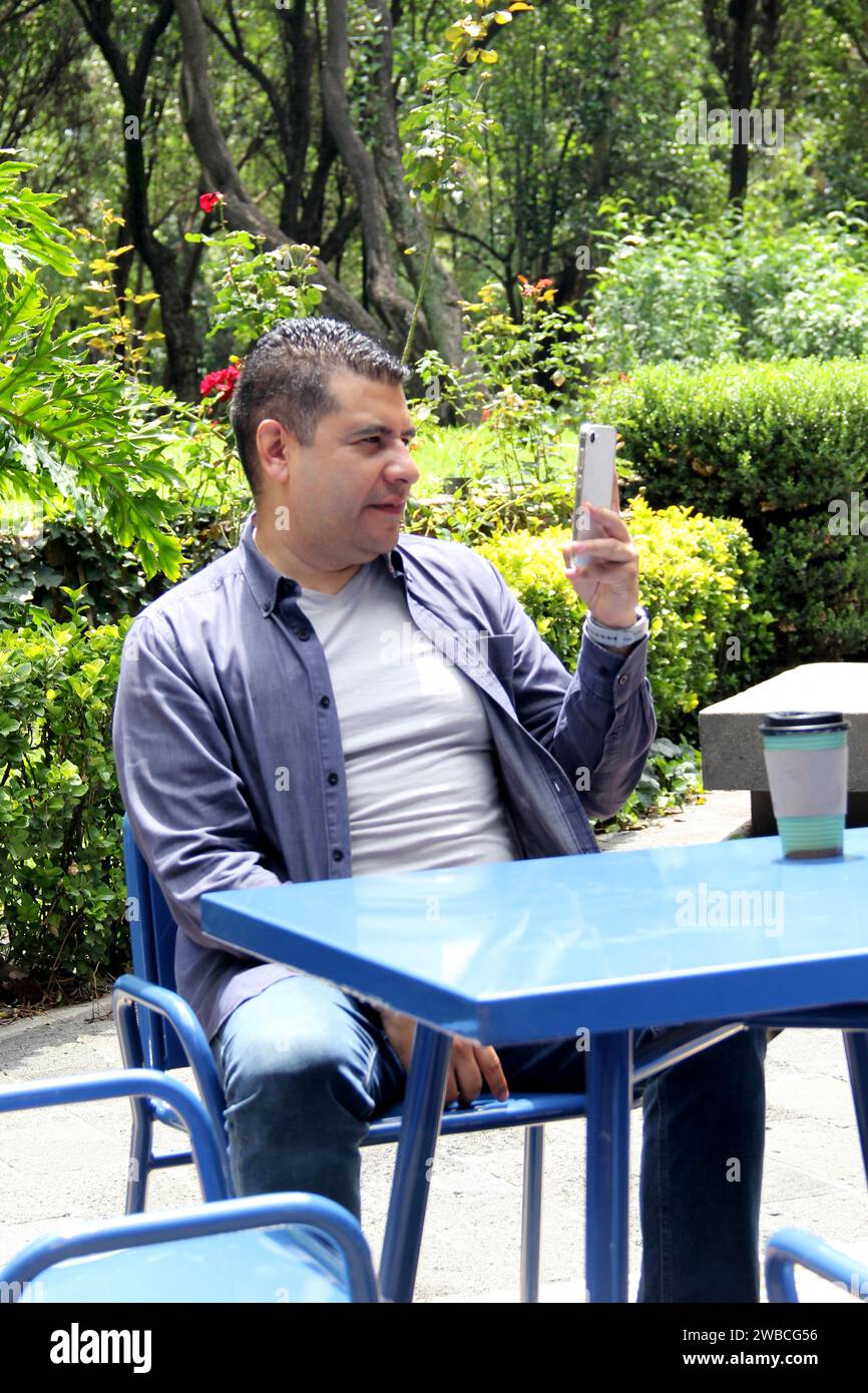 Un Latino de 40 ans à la peau foncée boit un café et utilise son téléphone portable sur un banc du parc pour travailler à distance, passer des appels, envoyer des messages, acheter et payer Banque D'Images