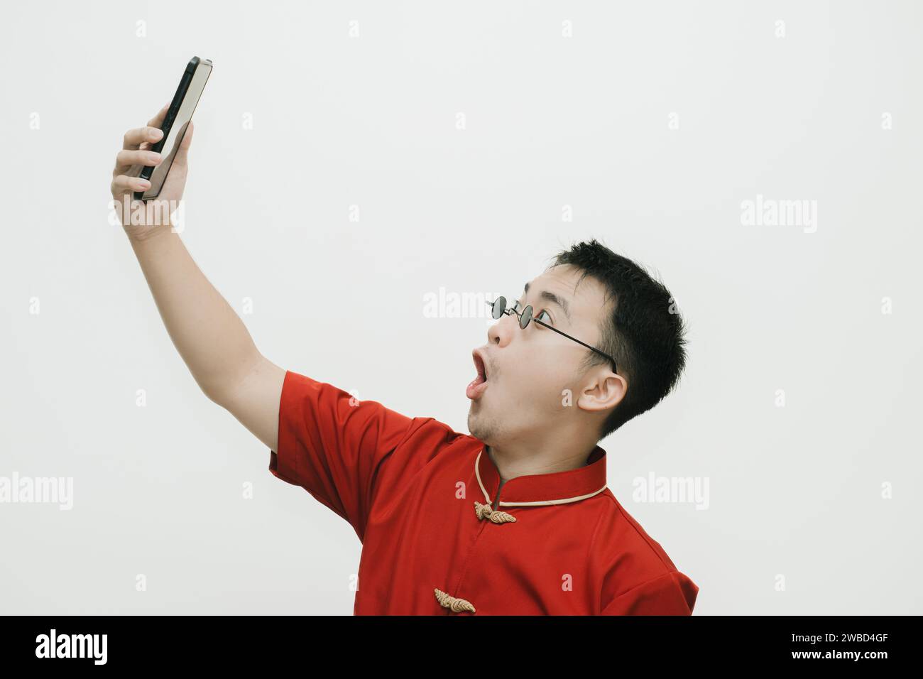 Visage choqué de l'homme asiatique portant Cheongsam et lunettes de soleil vintage noir tout en regardant le smartphone sur fond blanc Banque D'Images