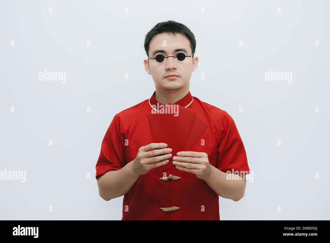 Homme asiatique portant Cheongsam et lunettes de soleil vintage rondes noires tenant angpao ou cadeau monétaire rouge et regardant l'appareil photo sur fond blanc Banque D'Images