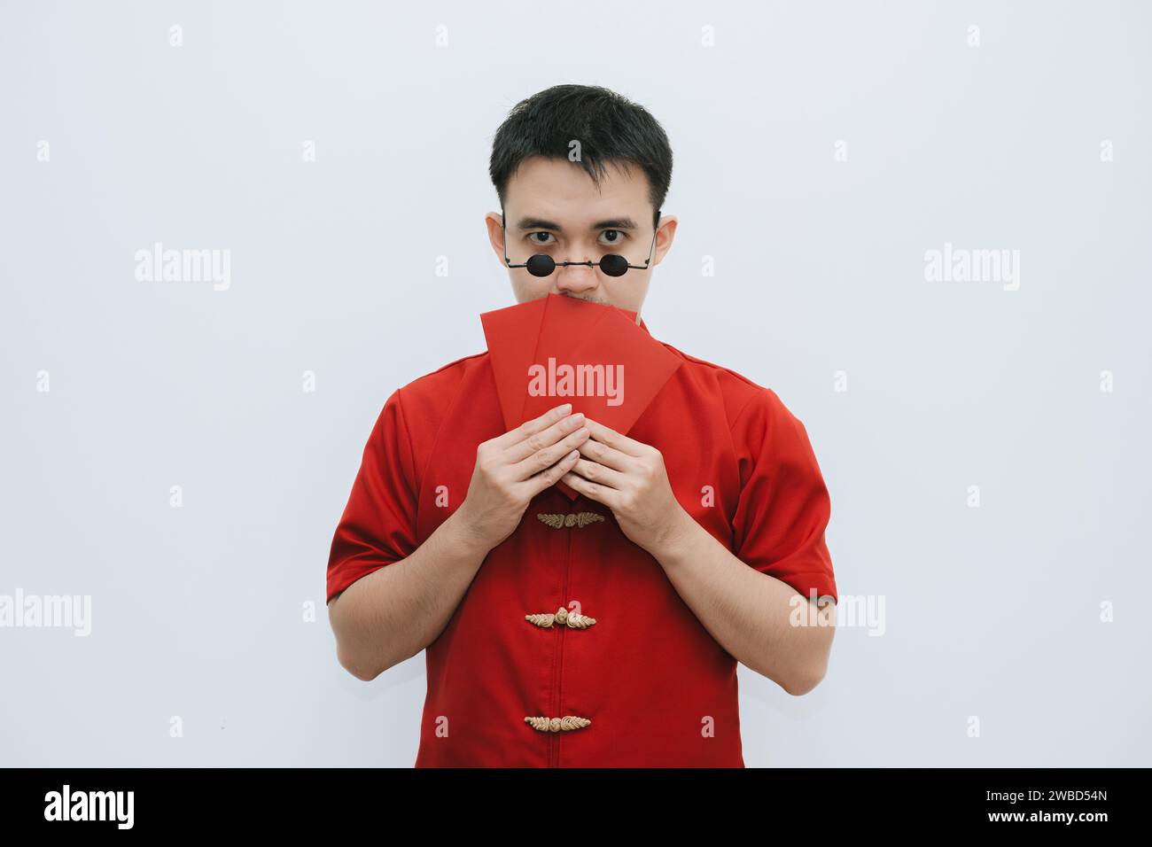 Homme asiatique portant Cheongsam et lunettes de soleil vintage rondes noires tout en tenant le cadeau monétaire rouge et couvrant son visage avec angpao sur bac blanc Banque D'Images