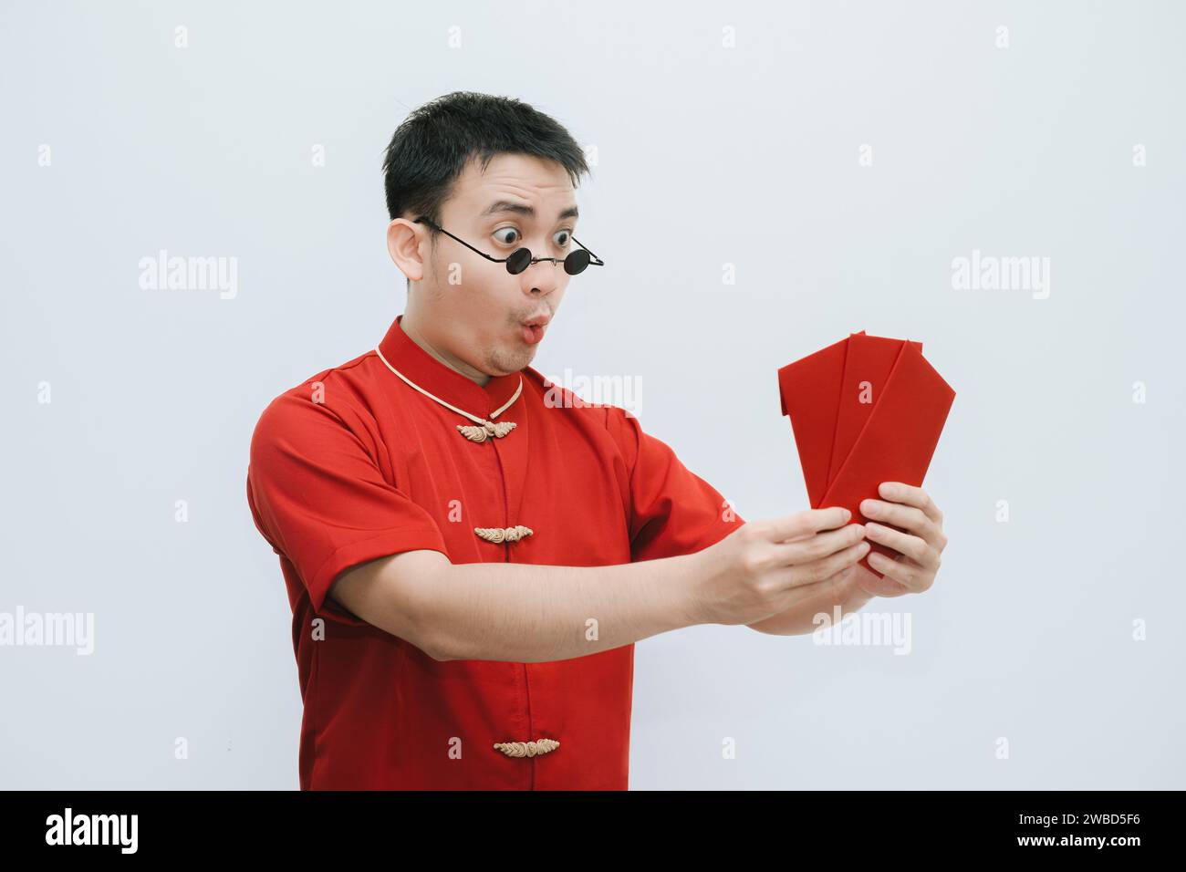 Visage choqué de l'homme asiatique portant Cheongsam et lunettes de soleil vintage rondes noires tout en tenant et en regardant l'angpao ou cadeau monétaire rouge sur blanc Banque D'Images