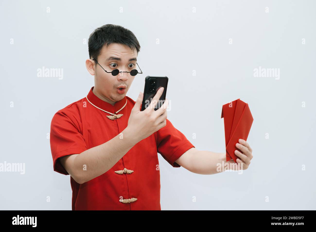 Visage choqué de l'homme asiatique portant Cheongsam et lunettes de soleil vintage rondes noires tout en tenant à l'angpao ou cadeau monétaire rouge et en regardant son s. Banque D'Images
