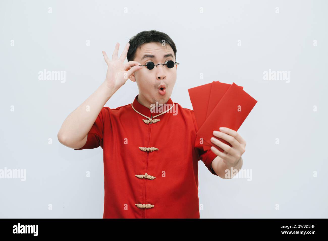 Visage choqué de l'homme asiatique portant Cheongsam et lunettes de soleil vintage rondes noires tout en tenant et en regardant l'angpao ou cadeau monétaire rouge sur blanc Banque D'Images