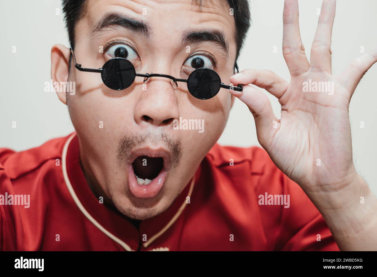Visage choqué de l'homme asiatique portant Cheongsam et lunettes de soleil vintage rondes noires en regardant l'appareil photo. Gros plan. Banque D'Images