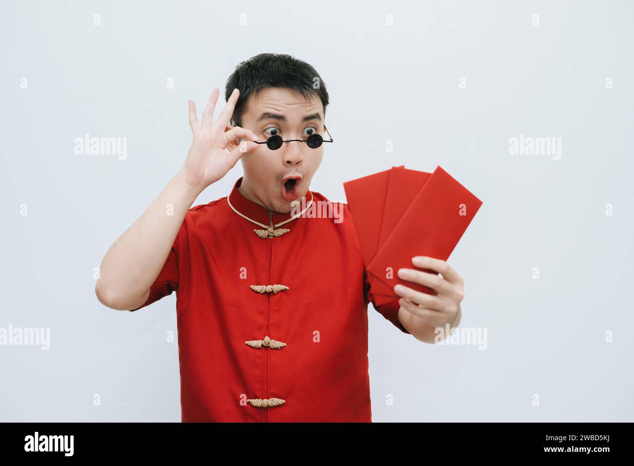 Wow visage de l'homme asiatique portant Cheongsam et lunettes de soleil vintage rondes noires tout en tenant et en regardant l'angpao ou cadeau monétaire rouge sur bac blanc Banque D'Images