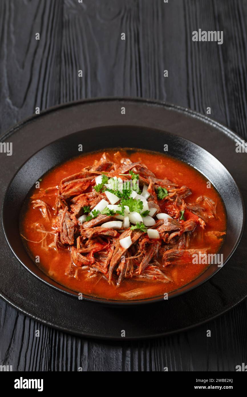 birria de res, ragoût de bœuf mexicain dans une sauce au piment rouge avec oignon cru et coriandre hachée dans un bol noir sur une table en bois noir, vue verticale, gratuit Banque D'Images