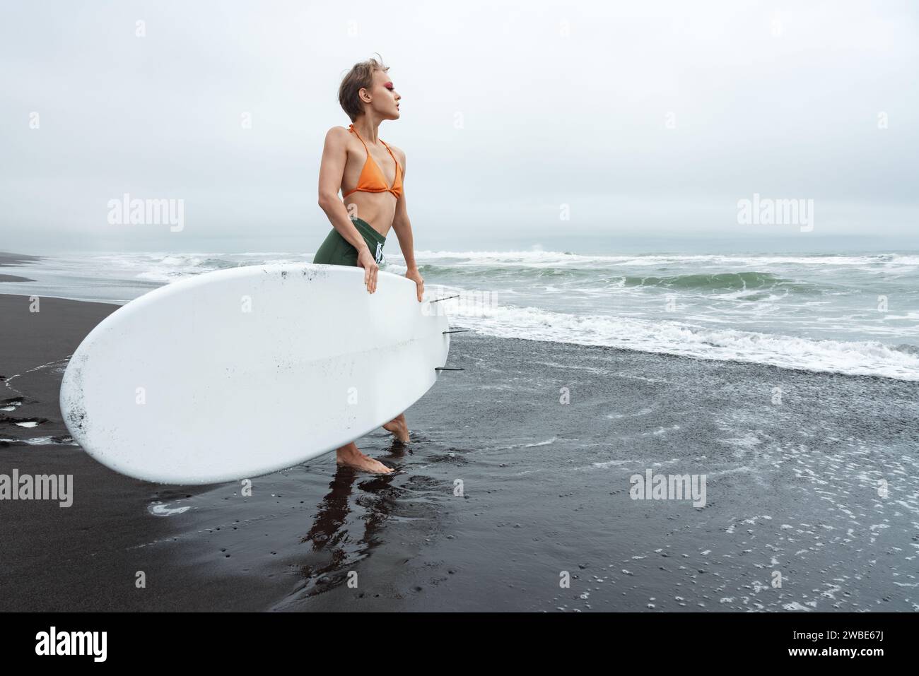 Séduisant surfeur féminin debout sur la plage de sable noir tenant la planche de surf blanche pendant les vacances d'été à la plage Banque D'Images