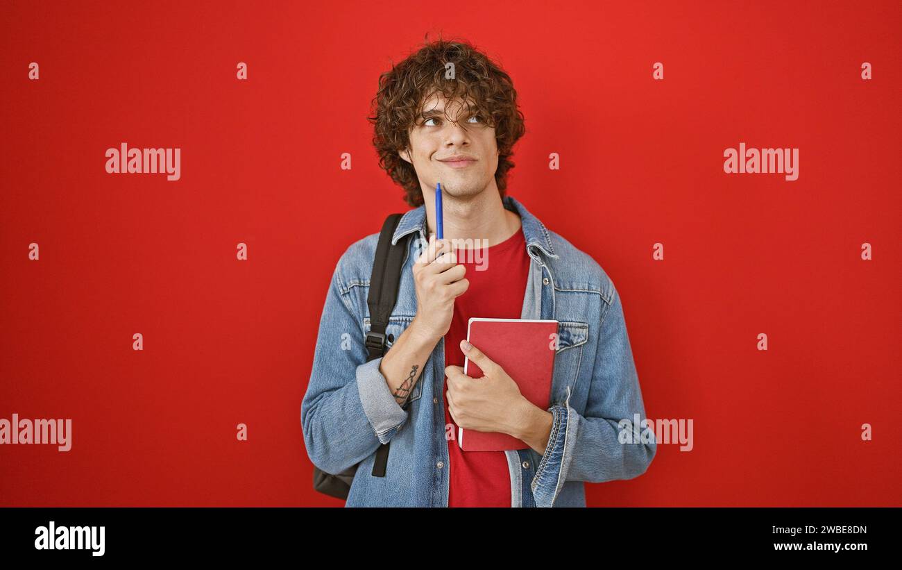 Un jeune homme réfléchi aux cheveux bouclés tient un livre sur fond rouge, évoquant une ambiance décontractée et contemplative. Banque D'Images