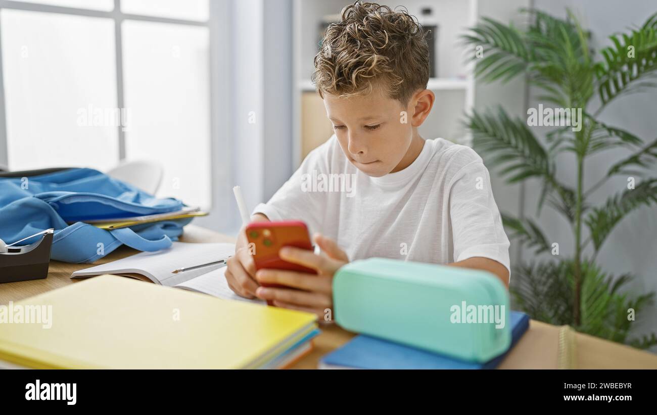 Adorable garçon blond étudiant, un cookie intelligent, prenant des notes sérieuses de son smartphone tout en regardant son écran dans la salle de classe Banque D'Images