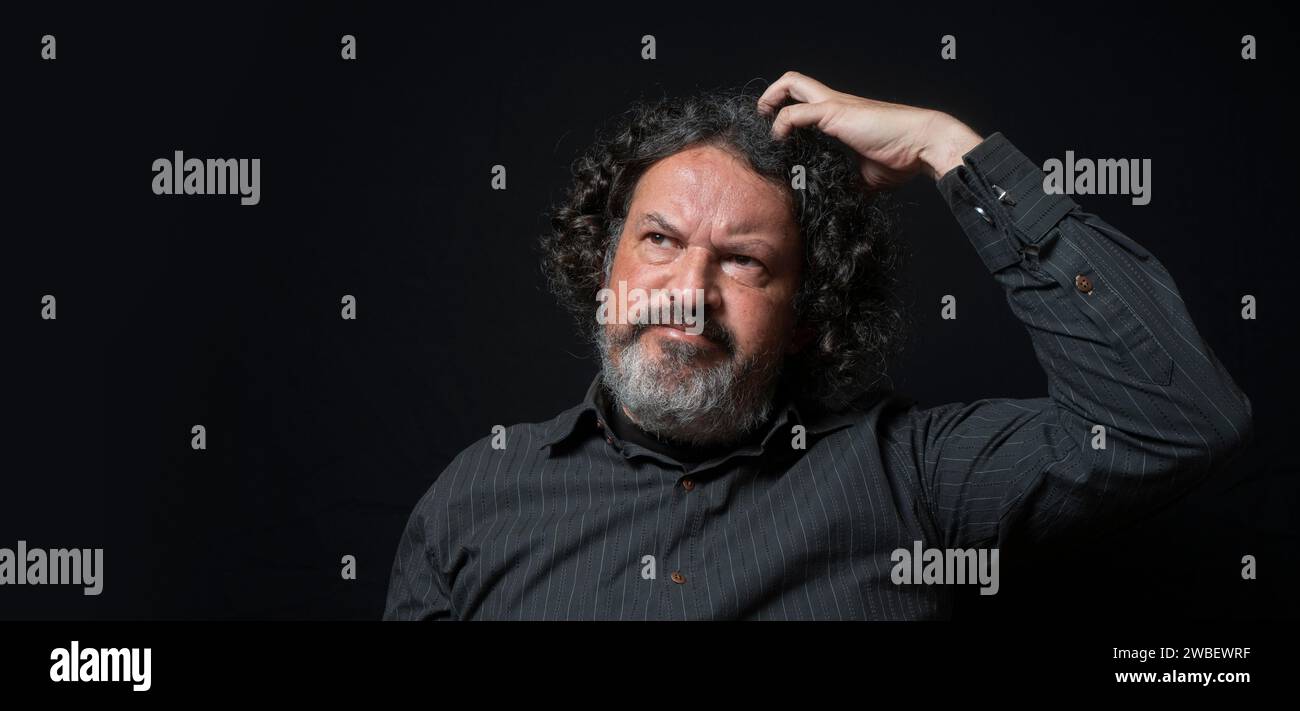 Portrait d'homme latin avec barbe blanche et cheveux bouclés noirs avec une expression douteuse, grattant la tête avec la main, portant une chemise noire contre bac noir Banque D'Images