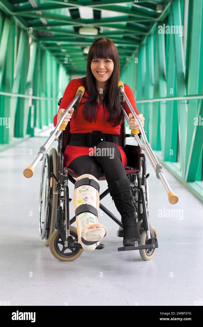 Une jeune femme avec une jambe en plâtre est assise dans un fauteuil roulant et sourit, à l'hôpital Banque D'Images