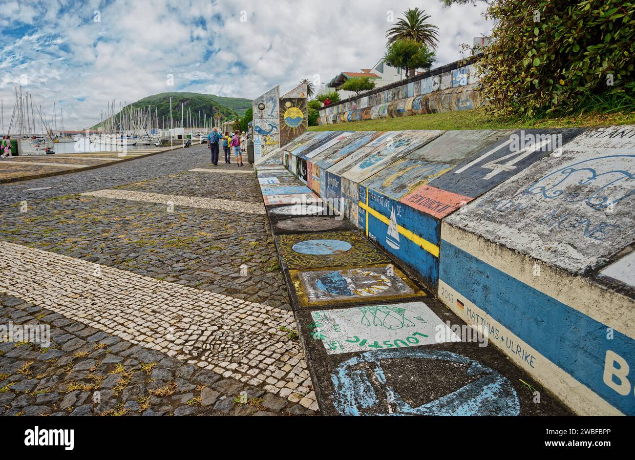 Art mural coloré des traversées de l'Atlantique le long d'un mur de port avec des marcheurs et des voiliers en arrière-plan, Horta, Faial Island, Açores, Portugal Banque D'Images