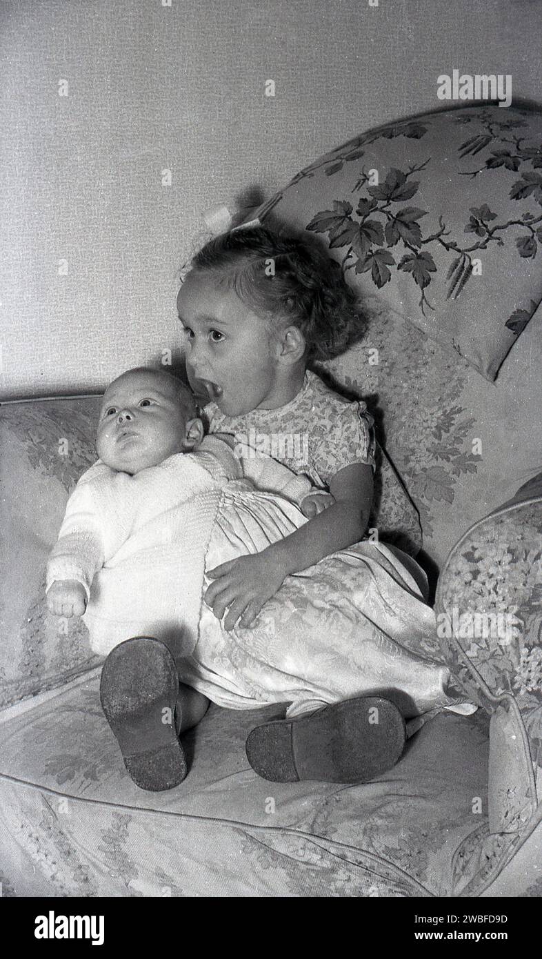 Années 1960, historique, une jeune fille assise sur une chaise longue avec sa nouvelle petite sœur sur ses genoux, Angleterre, Royaume-Uni. Banque D'Images