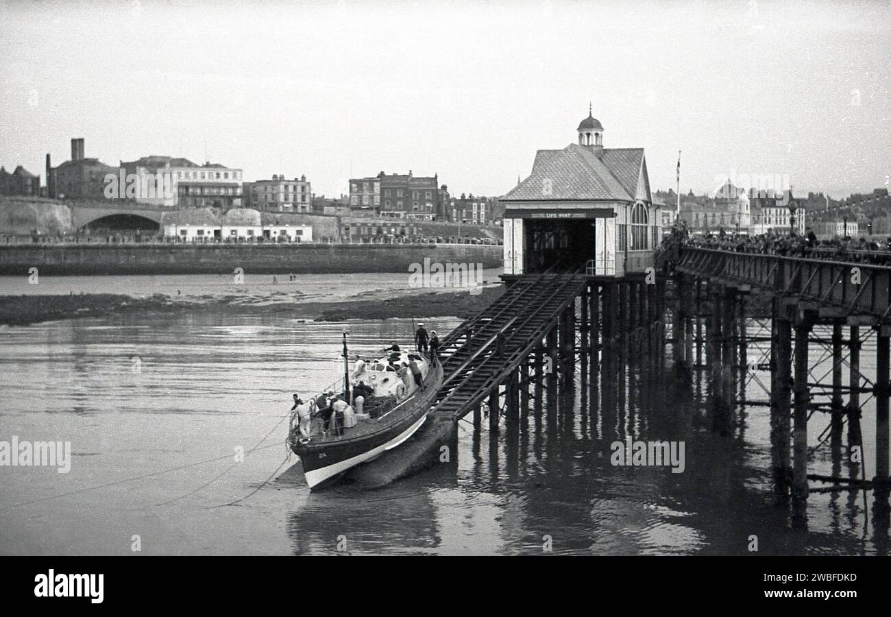 Années 1960, historique, station de bateau de sauvetage Margate, avec bateau de sauvetage sur la cale et les gens sur la passerelle de la jetée de fer victorienne à Margate, Kent, Angleterre, Royaume-Uni. Margate jetée ou jetée comme elle était connue, a été conçue par Eugenius Birch et a ouvert en 1855, remplaçant une jetée en bois antérieure. C'était la première jetée de bord de mer construite en fer dans le monde. Le hangar à bateaux et la cale de cale du bateau de sauvetage RNLI ont été ajoutés à la jetée en 1898 et ont fonctionné jusqu'en 1978, lorsqu'une grave tempête a détruit la majeure partie de la jetée qui avait déjà été fermée deux ans plus tôt pour des raisons de sécurité. Banque D'Images