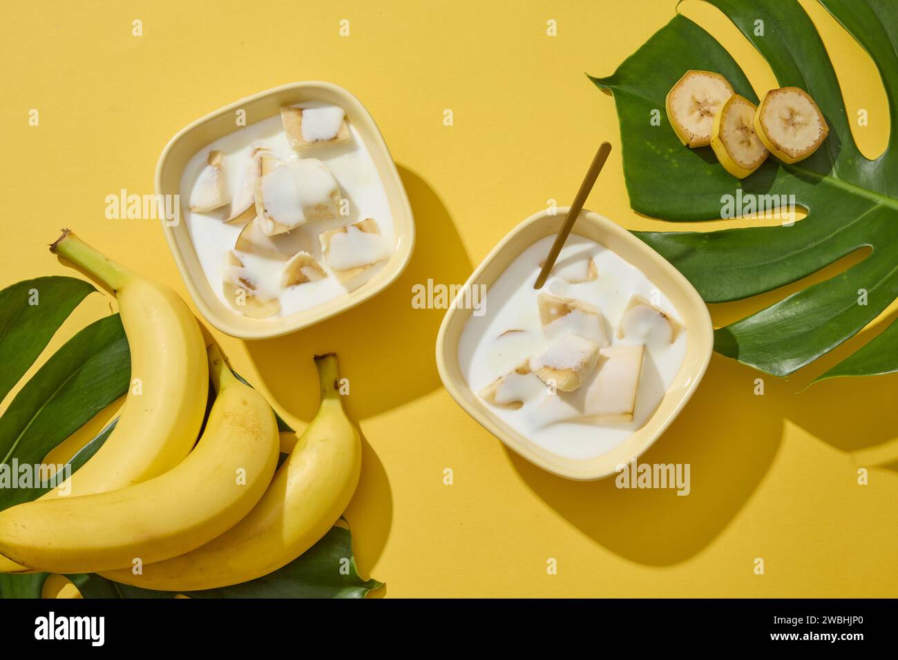 Vue de dessus de quelques bananes et bols de yaourt et tranches de banane. Fond jaune avec des feuilles tropicales. La banane (Musaceae) est un excellent hydratant naturel Banque D'Images