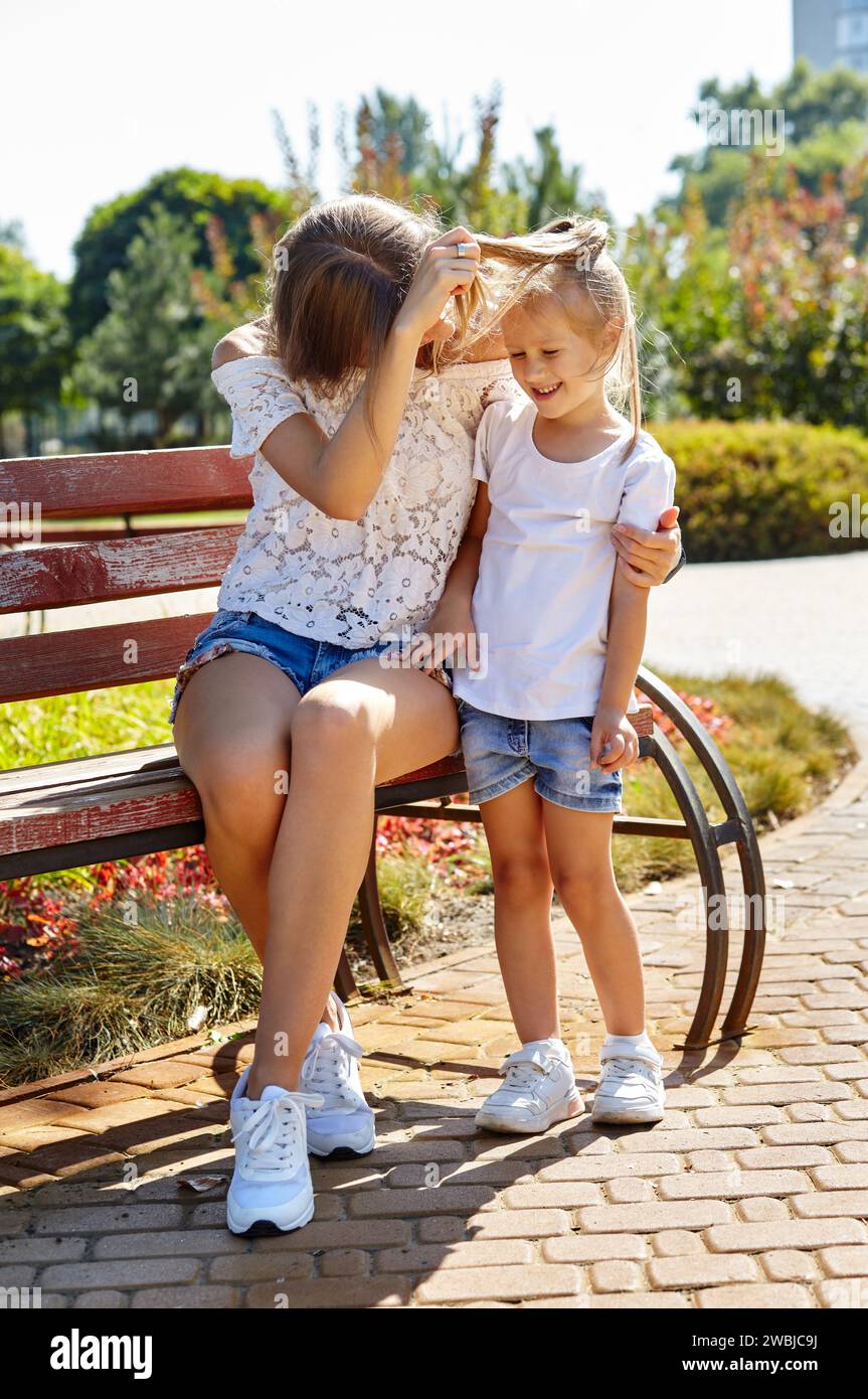Mère et fille assises sur un banc dans le parc ensoleillé de la ville d'été. Enfance, loisirs et concept de personnes - repos familial heureux et passer un bon moment Banque D'Images