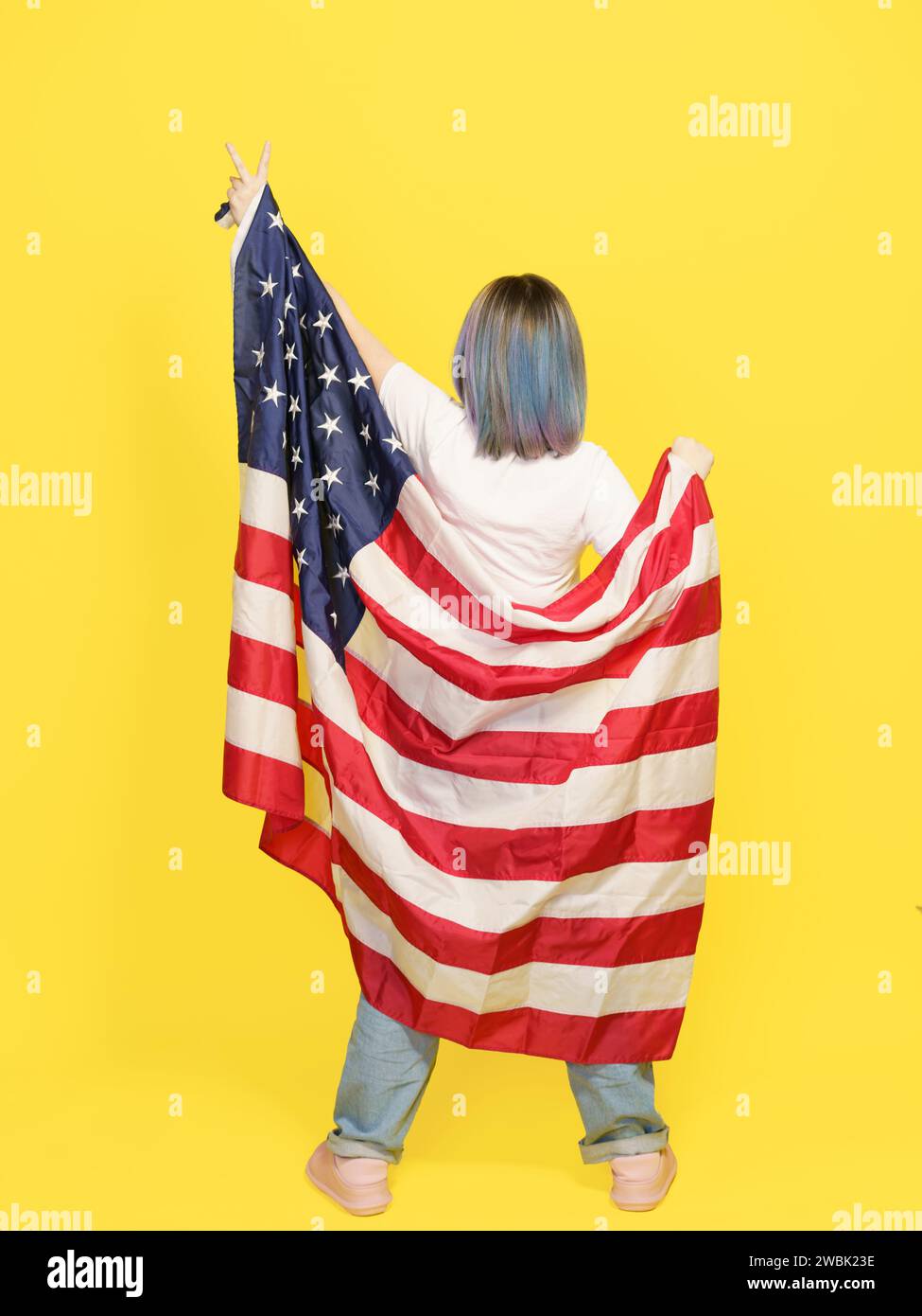 Signe de victoire par fille couvert de drapeau américain, fierté patriotique, diversité culturelle et liberté. Adolescent isolé sur fond jaune avec espace de copie pour votre texte. Vue arrière pleine hauteur . Photo de haute qualité Banque D'Images