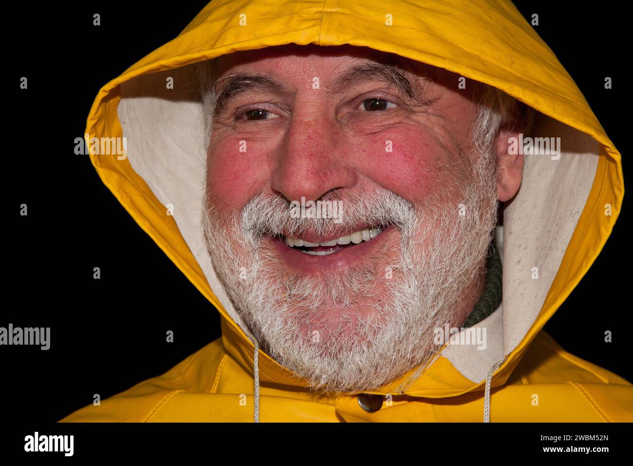 Le vieux pêcheur à la barbe frissonnée, à la veste de pluie jaune et à la capuche rit, tandis que les rides autour de ses yeux révèlent une histoire de vie pleine d'expériences. Banque D'Images