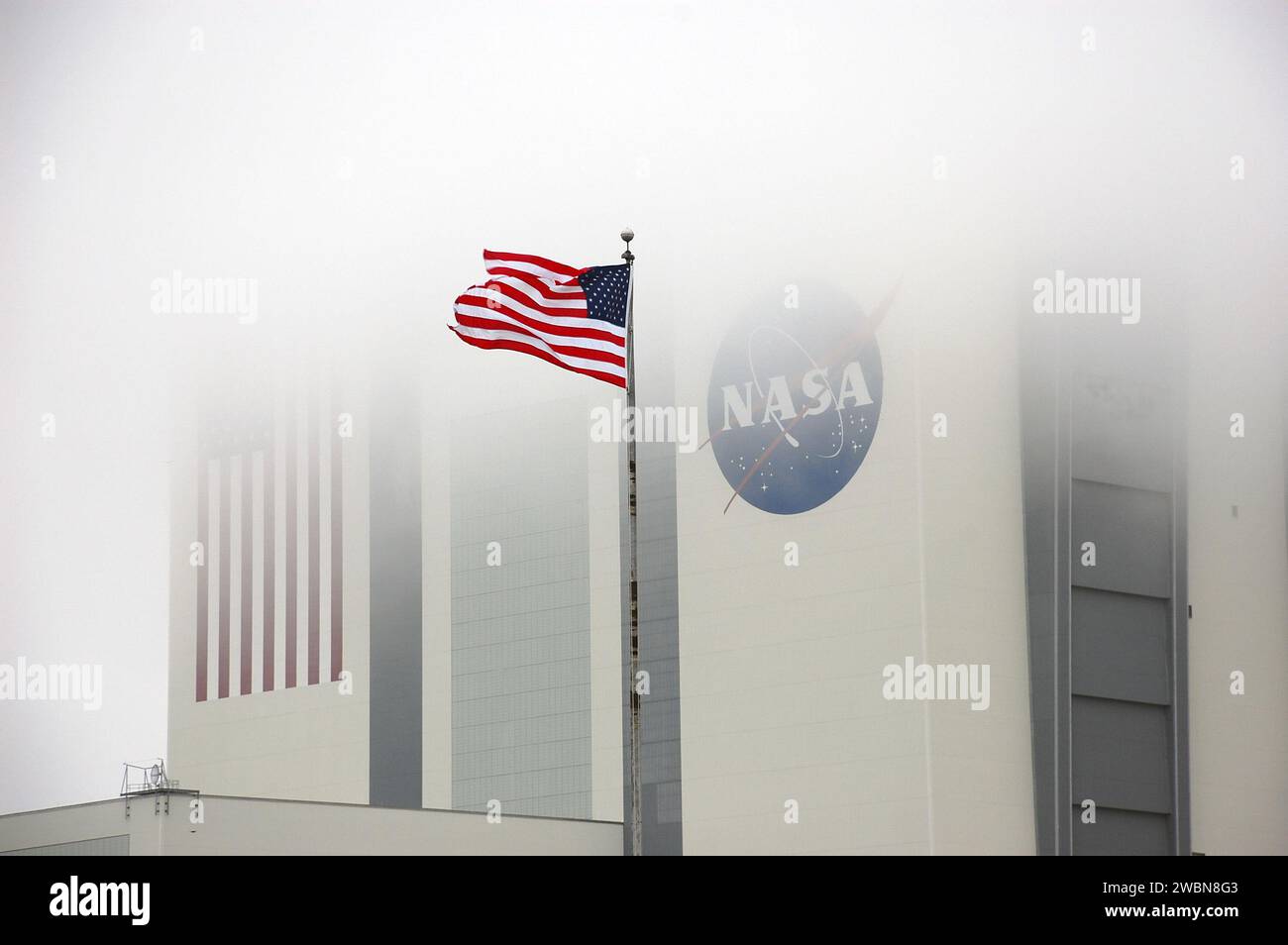 CAP CANAVERAL, Floride. – Au Kennedy Space Center de la NASA en Floride, un drapeau américain flotte fièrement près du bâtiment d'assemblage de véhicules de 525 pieds de haut, obscurci dans le brouillard. Le bâtiment massif, autrefois utilisé pour traiter les fusées Apollo Saturn V et les navettes spatiales pour le lancement, subit une transformation pour le préparer à supporter la fusée de prochaine génération de la NASA, le Space Launch System, ou SLS, ainsi que la myriade de fusées commerciales en cours de développement. Banque D'Images