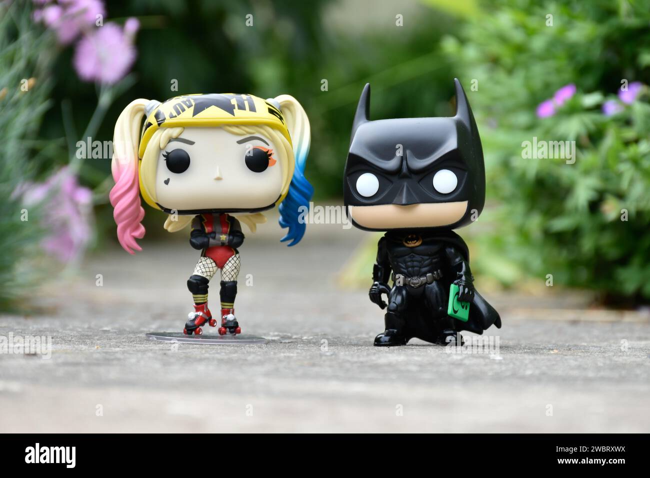 Funko Pop figurines d'action de DC comics super-héros Harley Quinn et Batman debout sur la route asphaltée dans le parc de printemps, herbe verte, fleurs roses. Banque D'Images