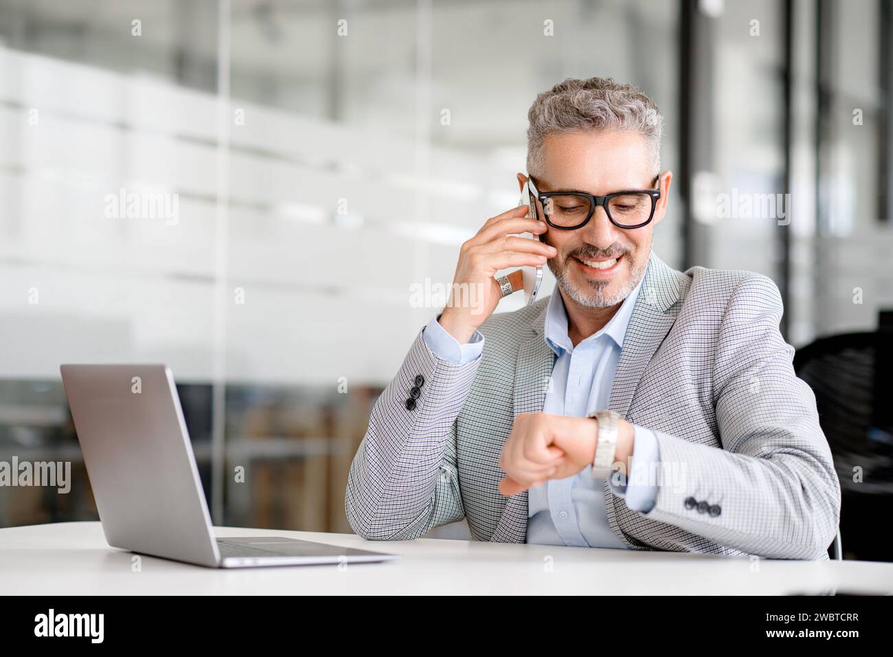 Homme d'affaires optimiste senior des années 60 appelant sur smartphone un regard sur montre-bracelet ou montre intelligente sur sa main assis dans le lieu de travail dans le bureau moderne. Ponctualité et concept de personnes Banque D'Images