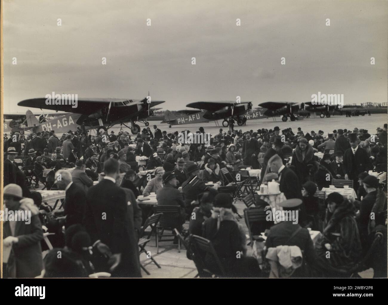 Événement aéronautique, vraisemblablement Schiphol, c. 1925 - c. 1935 Photography Album Magazine avec une photo de personnes assises sur des tables sur un aéroport avec quatre avions stationnés en arrière-plan. Partie de l'album commémoratif de la ligne Indes de KLM, partie I. Journal Schiphol. support photographique. carton gélatine argent impression trafic aérien, aviation. voyages ; tourisme. Aérodrome, aéroport Schiphol Banque D'Images