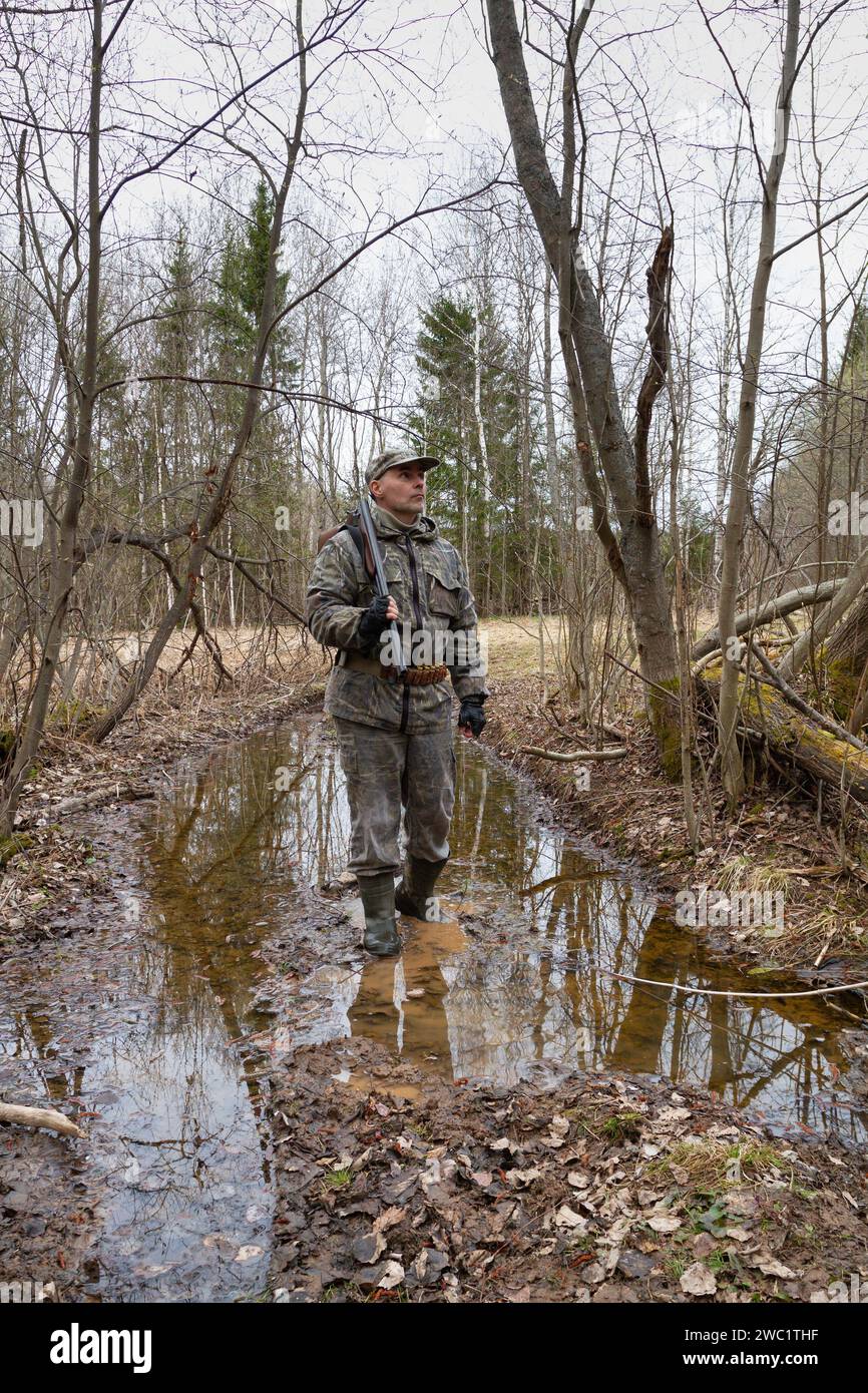 Un chasseur se fraie un chemin à travers une flaque d'eau dans la forêt printanière. Il a un fusil de chasse pendu sur son épaule. Le crépuscule du soir s'approfondit. Banque D'Images