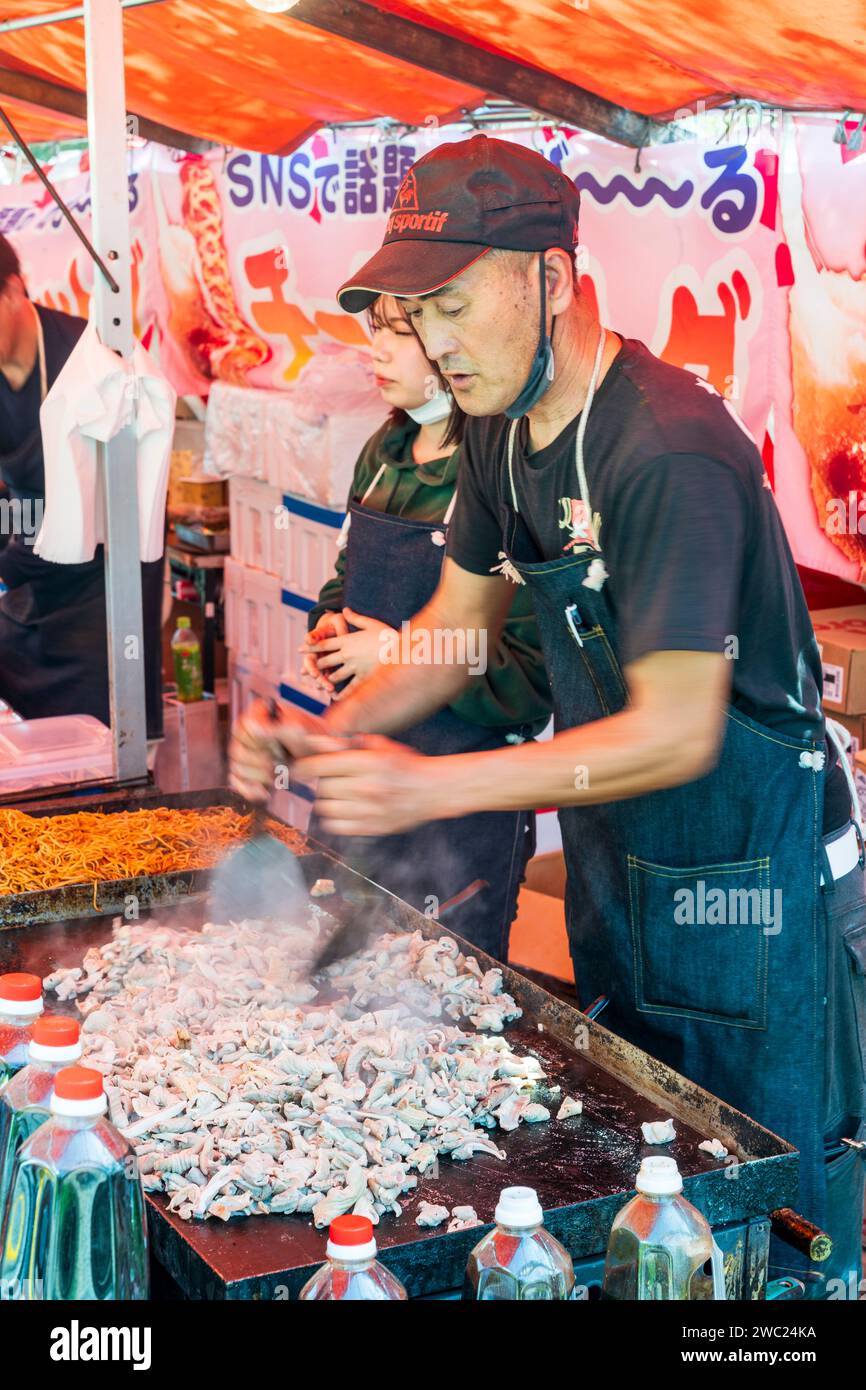 Stalle de restauration rapide au Japon pendant le festival printanier des cerisiers en fleurs. Homme utilisant deux spatules métalliques pour préparer des aliments sur une plaque chauffante à côté d'un plateau de nouilles. Banque D'Images