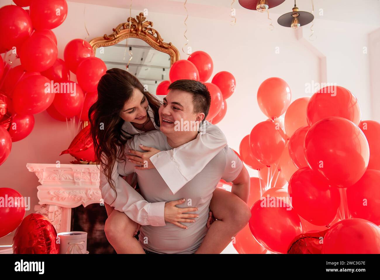 Moment ludique et romantique entre couple fou près de ballons rouges dans une pièce lumineuse aux intérieurs blancs. Femme assise sur les hommes dos. Couple qui se trompe Banque D'Images