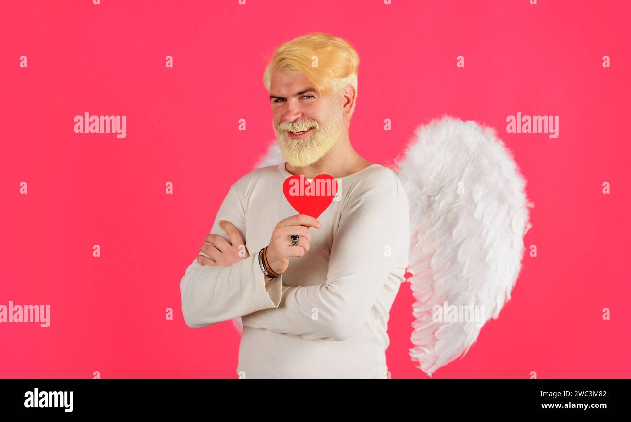 Fête de la Saint-Valentin. Homme barbu souriant dans des ailes d'ange avec carte de Saint-Valentin. Ange cupidon mâle avec coeur de papier rouge, symbole de l'amour et Banque D'Images