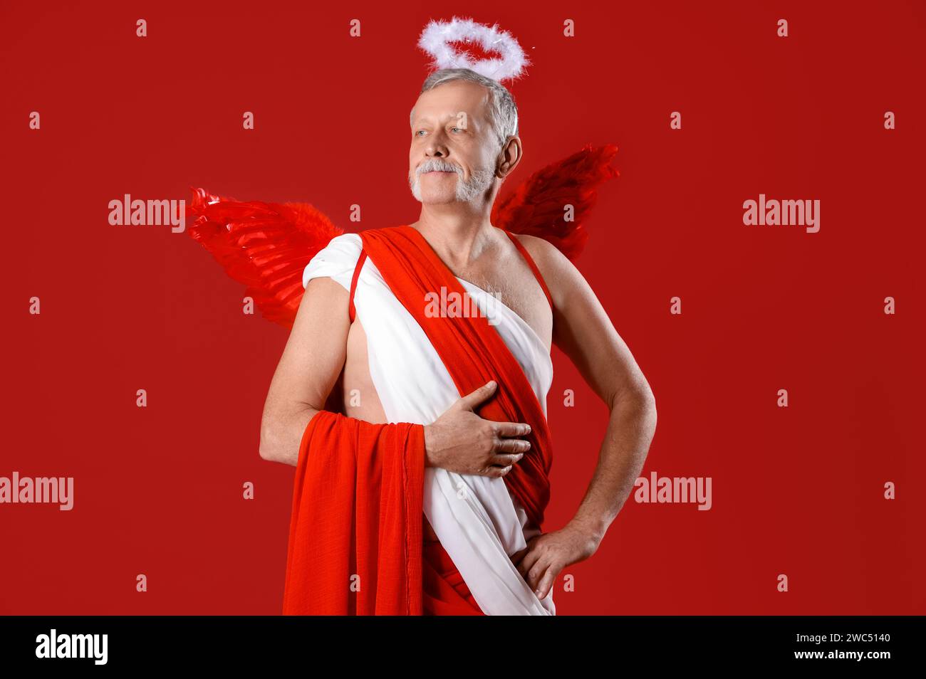 Homme mature habillé comme Cupidon sur fond rouge. Fête de la Saint-Valentin Banque D'Images