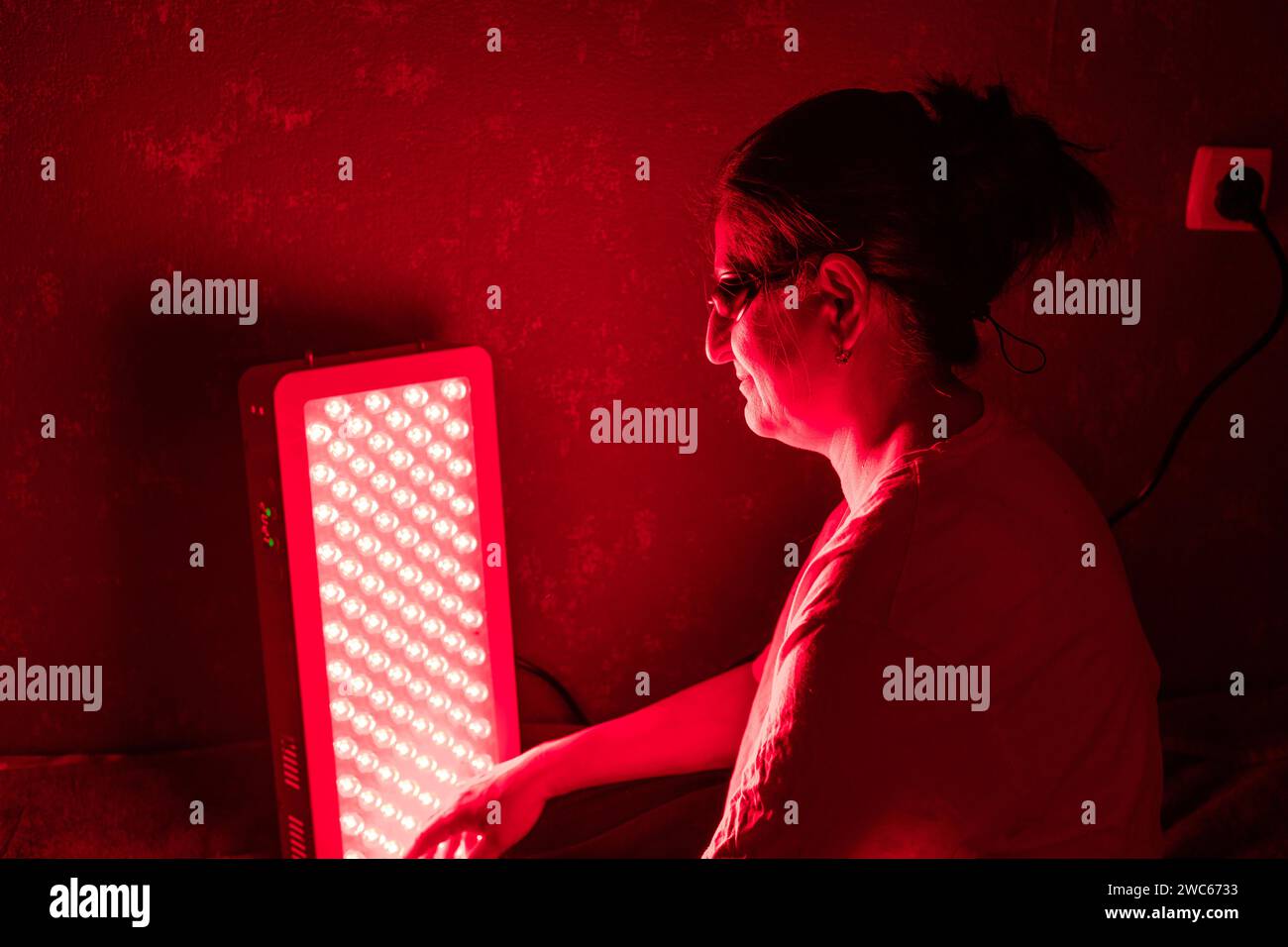 Femme âgée faisant la séance de thérapie de lumière rouge avec le panneau et portant une protection oculaire à la maison sur le lit Banque D'Images