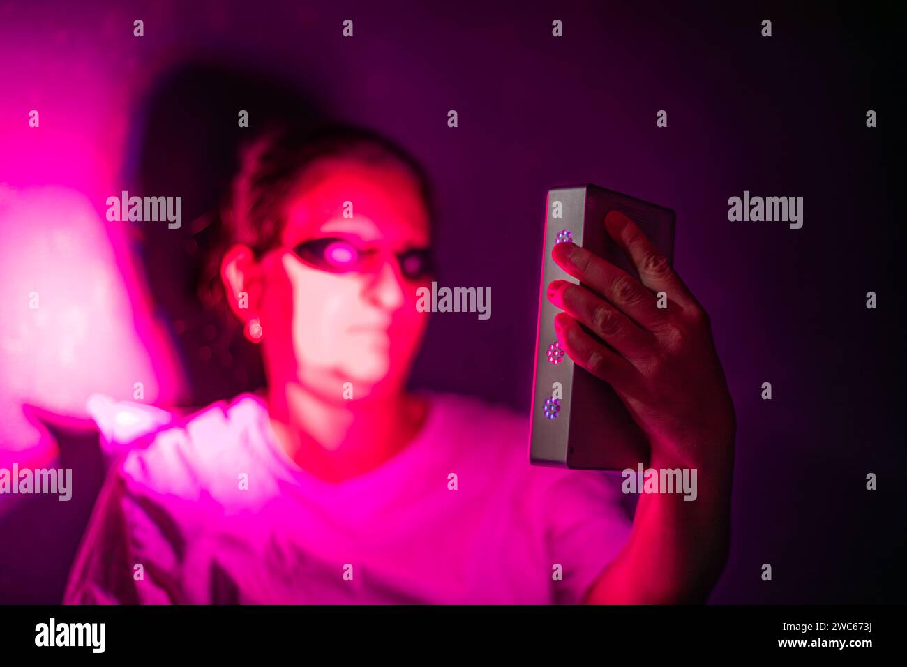 Gros plan d'une femme âgée faisant la séance de thérapie à la lumière rouge avec un appareil portatif et portant une protection oculaire à la maison sur le lit Banque D'Images