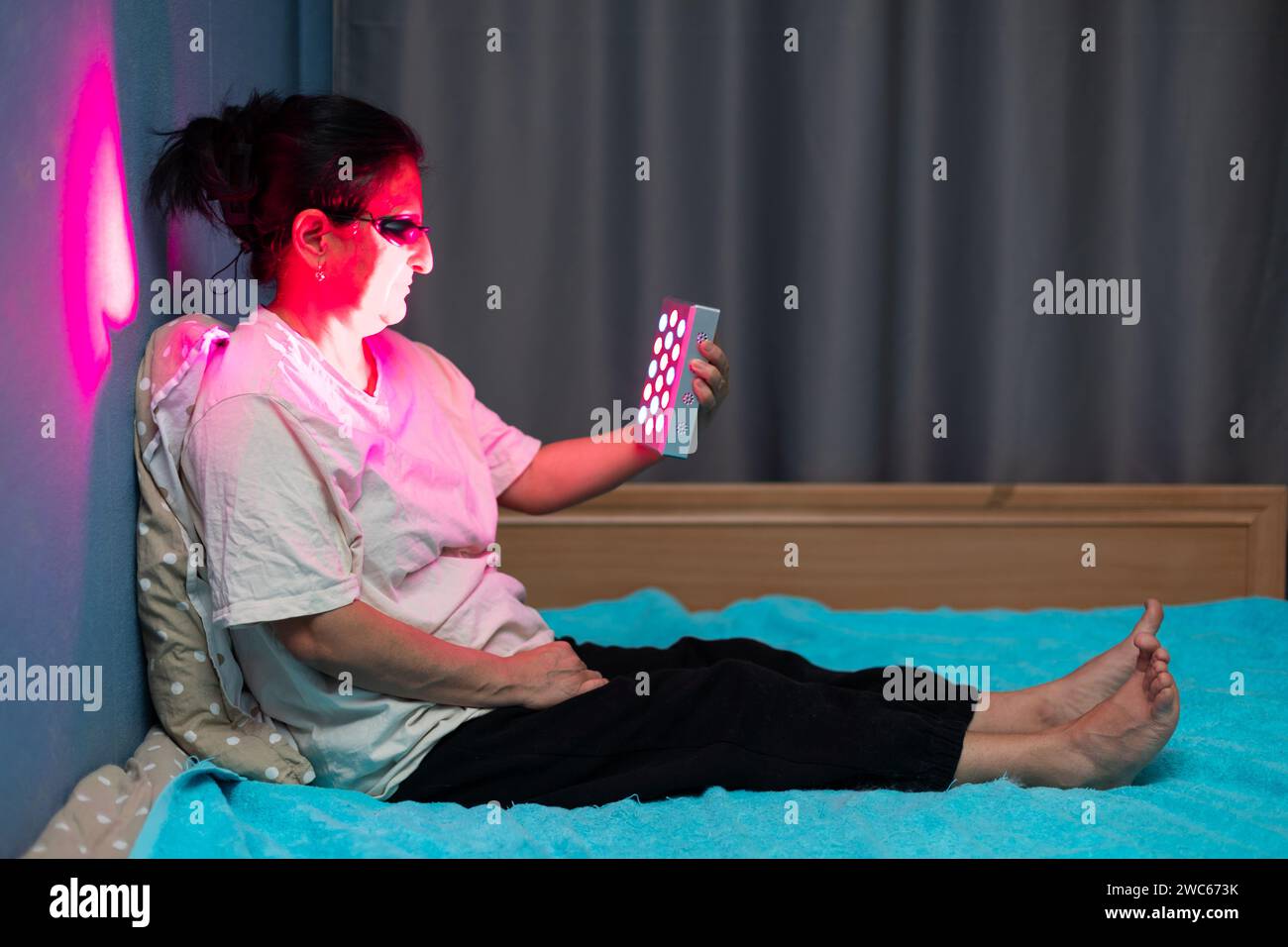 Femme âgée faisant la séance de thérapie à la lumière rouge avec un appareil portatif et portant une protection oculaire à la maison sur le lit Banque D'Images