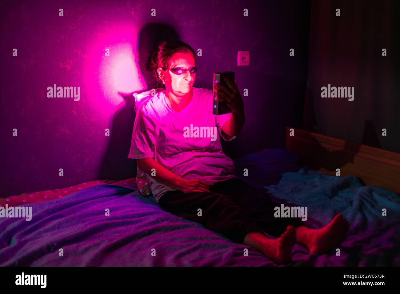 Femme âgée faisant la séance de thérapie à la lumière rouge avec un appareil portatif et portant une protection oculaire à la maison sur le lit Banque D'Images
