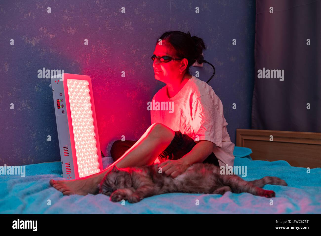 Femme âgée traitant la jambe douloureuse avec le panneau de thérapie de lumière rouge et portant une protection oculaire tout en caressant un chat à la maison sur le lit Banque D'Images