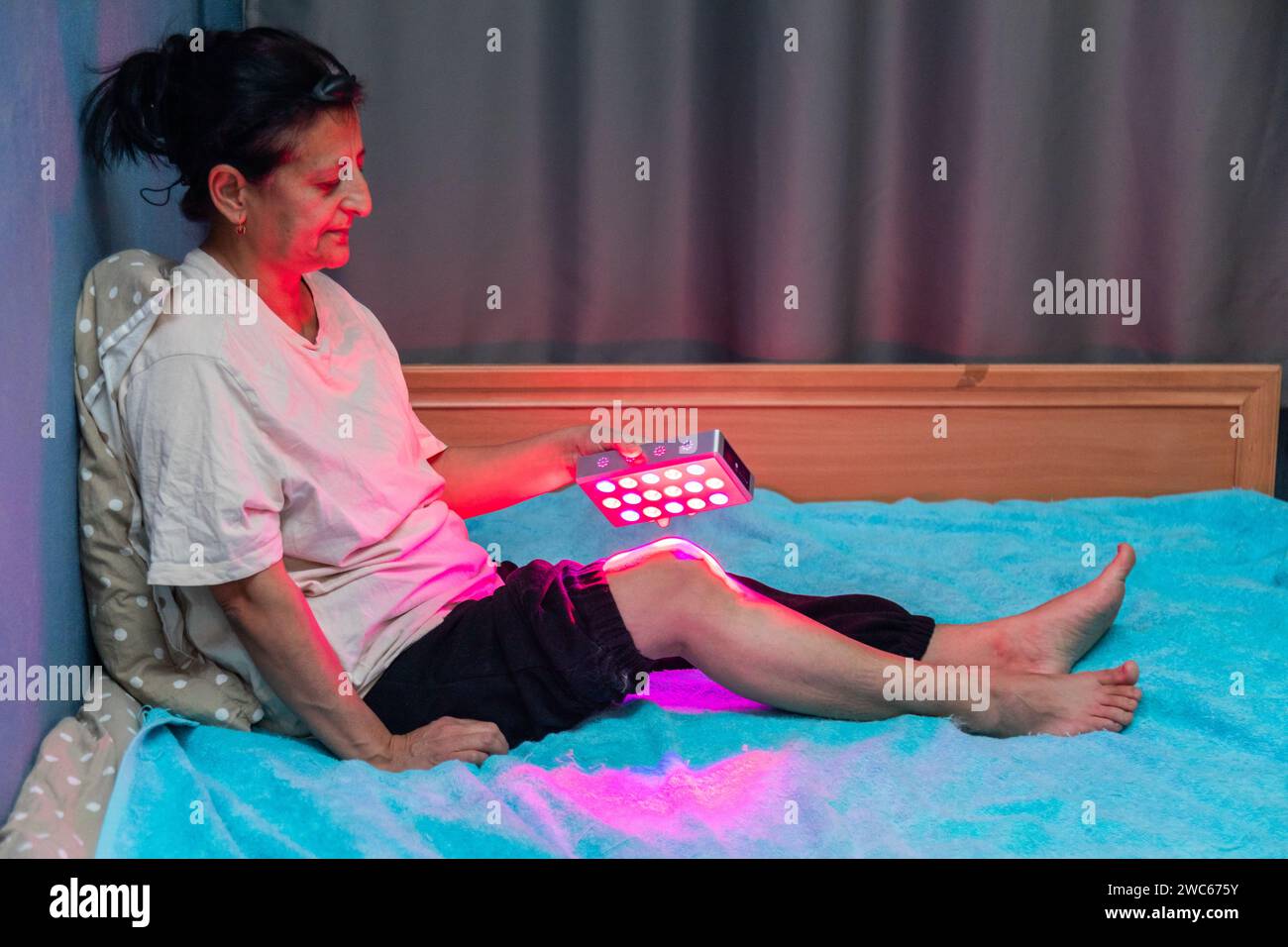 Femme âgée traitant une jambe douloureuse avec l'appareil portable de thérapie à la lumière rouge à la maison sur le lit Banque D'Images