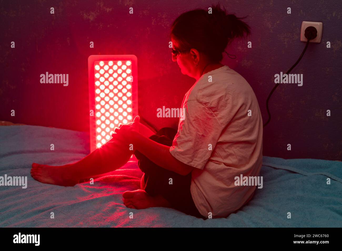 Femme âgée traitant la jambe douloureuse avec le panneau de thérapie de lumière rouge et portant une protection oculaire à la maison sur le lit Banque D'Images