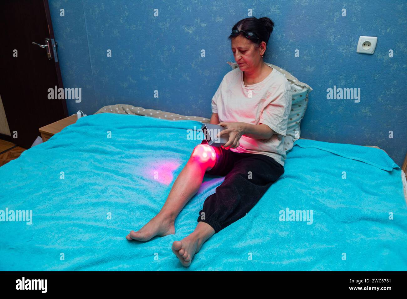 Femme âgée traitant une jambe douloureuse avec l'appareil portable de thérapie à la lumière rouge à la maison sur le lit Banque D'Images