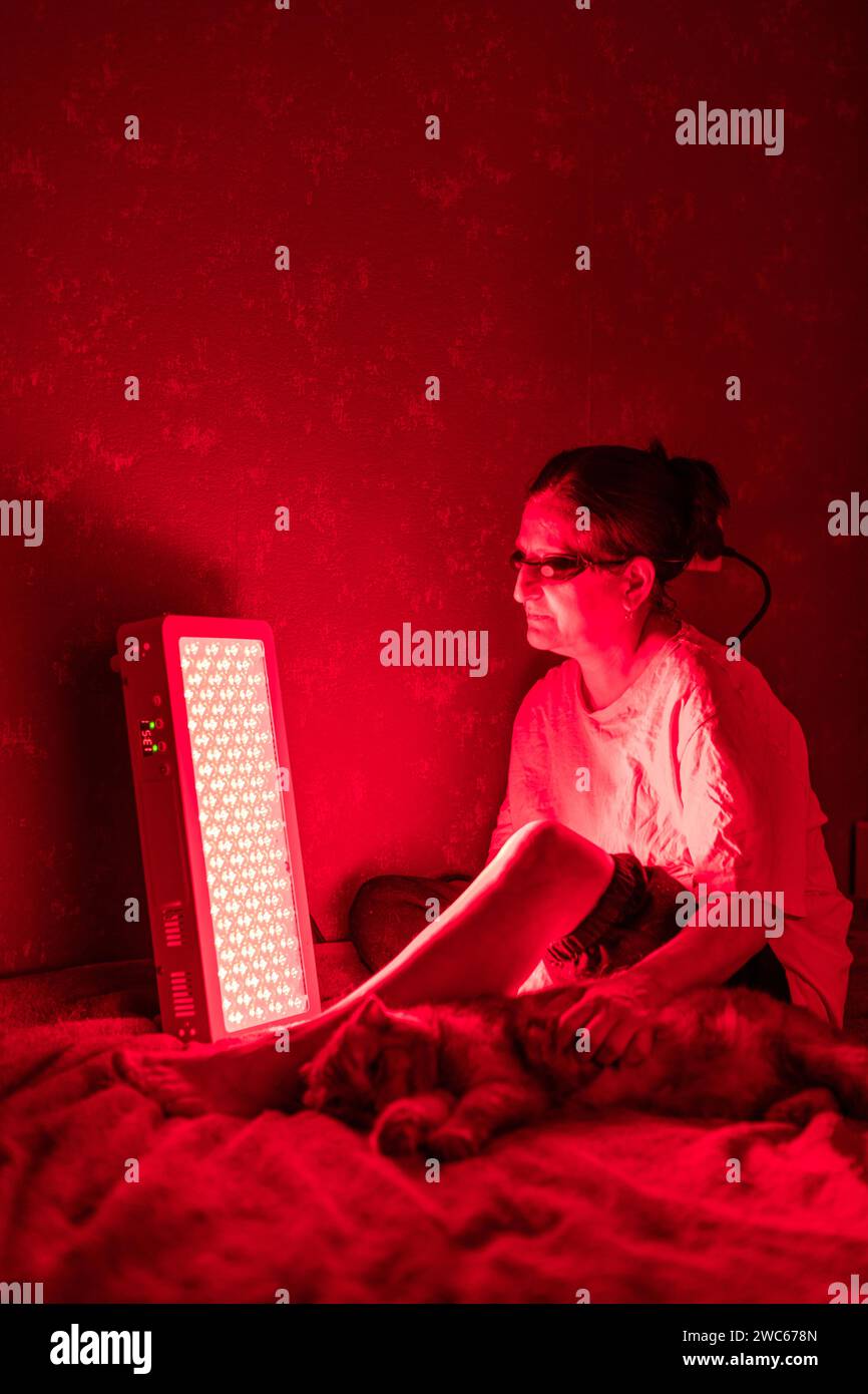 Femme âgée traitant la jambe douloureuse avec le panneau de thérapie de lumière rouge et portant une protection oculaire tout en caressant un chat à la maison sur le lit Banque D'Images
